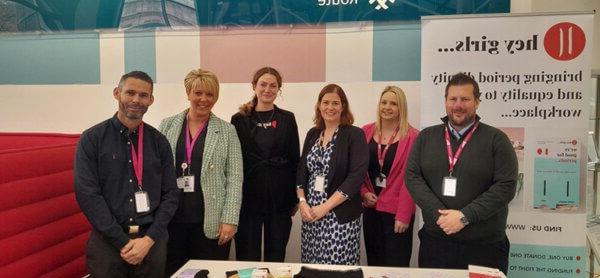 Mitie staff members at a Hey Girls stand - the pull-up banner behind says 'Bringing period dignity and equality to your workplace'